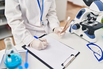 Young blonde woman wearing scientist uniform holding test tubes writing on checklist at laboratory