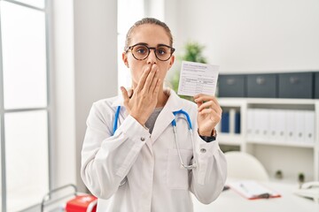 Young doctor woman holding covid certificate covering mouth with hand, shocked and afraid for...