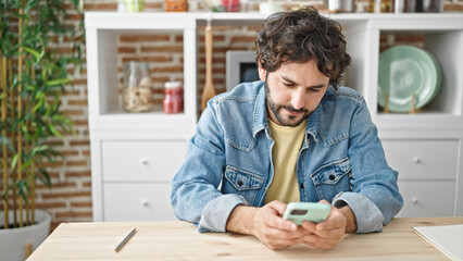 Young hispanic man using smartphone sitting on table at dinning room