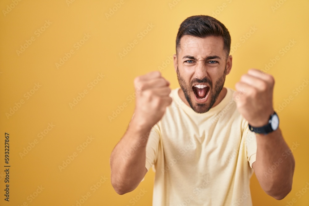 Sticker Handsome hispanic man standing over yellow background angry and mad raising fists frustrated and furious while shouting with anger. rage and aggressive concept.