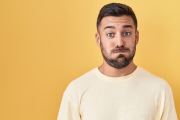Handsome hispanic man standing over yellow background puffing cheeks with funny face. mouth inflated with air, crazy expression. - obrazy, fototapety, plakaty