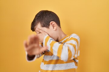 Young caucasian kid standing over yellow background covering eyes with hands and doing stop gesture...