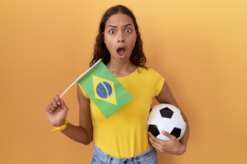 Young hispanic woman holding brazil flag and football ball in shock face, looking skeptical and sarcastic, surprised with open mouth