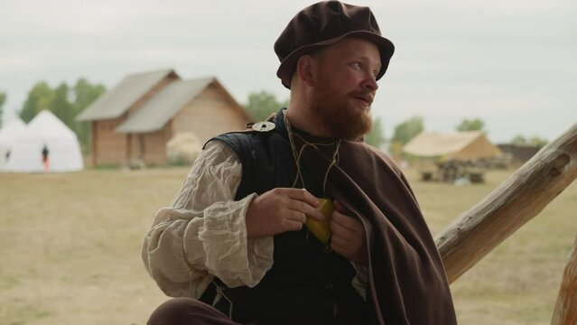 Bearded man in medieval clothing talks standing in yard in countryside. Village squire holds yellow potion bottle in bosom closeup on blurred background