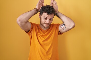Young hispanic man standing over yellow background doing bunny ears gesture with hands palms...