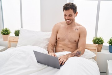 Young hispanic man using laptop sitting on bed at bedroom