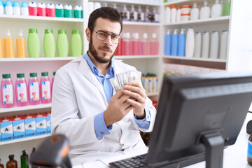 Young hispanic man pharmacist holding dollars using computer at pharmacy
