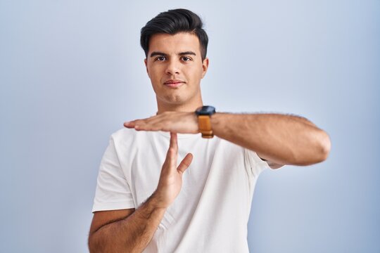 Hispanic man standing over blue background doing time out gesture with hands, frustrated and serious face