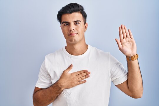 Hispanic man standing over blue background swearing with hand on chest and open palm, making a loyalty promise oath