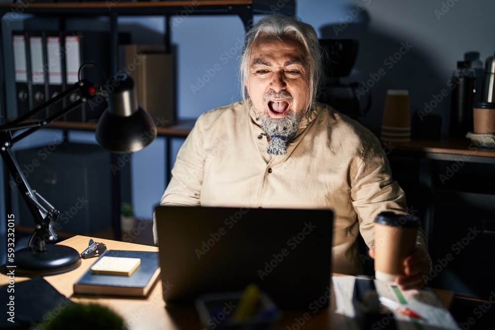 Sticker Middle age man with grey hair working at the office at night angry and mad screaming frustrated and furious, shouting with anger. rage and aggressive concept.