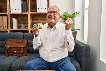 Senior therapist with grey hair playing colorful puzzle cube intelligence game screaming proud, celebrating victory and success very excited with raised arm
