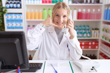Young caucasian woman working at pharmacy drugstore speaking on the telephone smiling doing phone...
