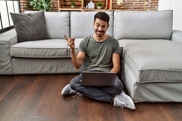 Young hispanic man using laptop sitting on the at home smiling happy pointing with hand and finger to the side