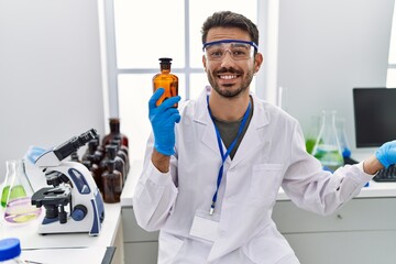 Young hispanic man working at scientist laboratory holding bottle celebrating achievement with happy smile and winner expression with raised hand