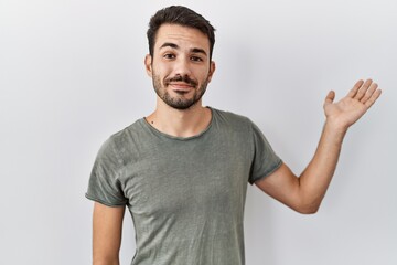 Young hispanic man with beard wearing casual t shirt over white background smiling cheerful presenting and pointing with palm of hand looking at the camera.
