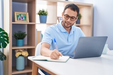 Young man using laptop writing on notebook at home