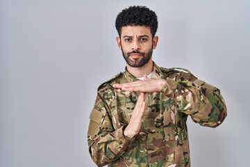 Arab man wearing camouflage army uniform doing time out gesture with hands, frustrated and serious face