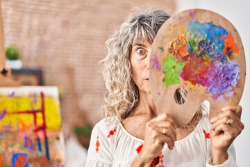 Fototapeta premium Middle age woman holding painter palette covering face clueless and confused expression. doubt concept.