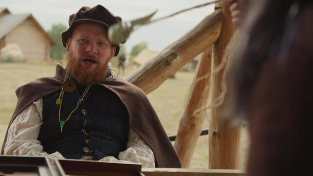 Red-bearded squire talks to woman sitting at table in courtyard in medieval village. Female hand spins pendant on rope in circle on blurred background closeup