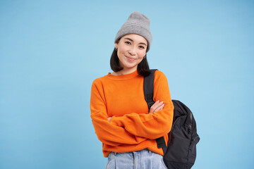 Cheerful chinese woman in warm hat and orange sweater, goes for walk with backpack, cross arms on...