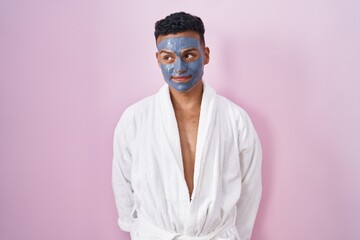 Young hispanic man wearing beauty face mask and bath robe smiling looking to the side and staring...