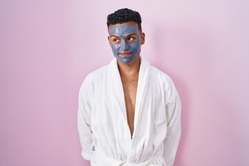 Young hispanic man wearing beauty face mask and bath robe smiling looking to the side and staring away thinking.