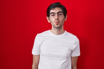 Obraz premium Young hispanic man standing over red background looking at the camera blowing a kiss on air being lovely and sexy. love expression.