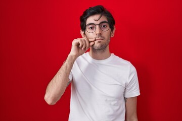 Young hispanic man standing over red background mouth and lips shut as zip with fingers. secret and...