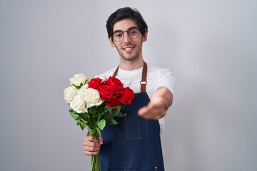 Young hispanic man holding bouquet of white and red roses smiling cheerful offering palm hand giving assistance and acceptance.