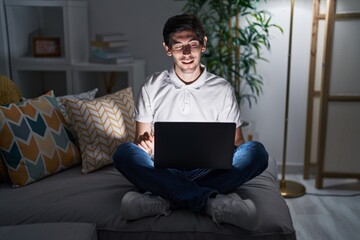 Young hispanic man using laptop at home at night winking looking at the camera with sexy expression, cheerful and happy face.