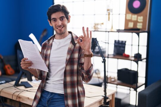 Young hispanic man reading music sheet at music studio doing ok sign with fingers, smiling friendly gesturing excellent symbol