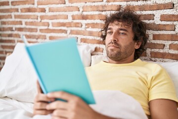 Young hispanic man reading book lying on bed at bedroom