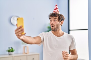 Hispanic young man wearing birthday hat doing video call with smartphone scared and amazed with open mouth for surprise, disbelief face