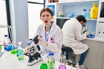 Young two people working at scientist laboratory scared and amazed with open mouth for surprise, disbelief face