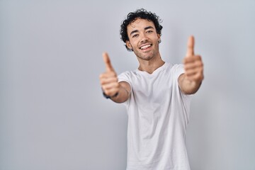 Hispanic man standing over isolated background approving doing positive gesture with hand, thumbs up smiling and happy for success. winner gesture.