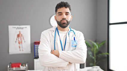 Young hispanic man doctor standing with serious expression and arms crossed gesture at clinic