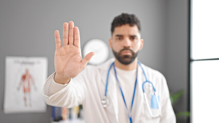 Young hispanic man doctor doing stop sign with hand at clinic