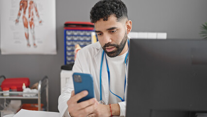 Young hispanic man doctor using smartphone and computer at clinic