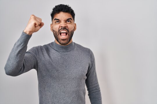 Hispanic man with beard standing over white background angry and mad raising fist frustrated and furious while shouting with anger. rage and aggressive concept.