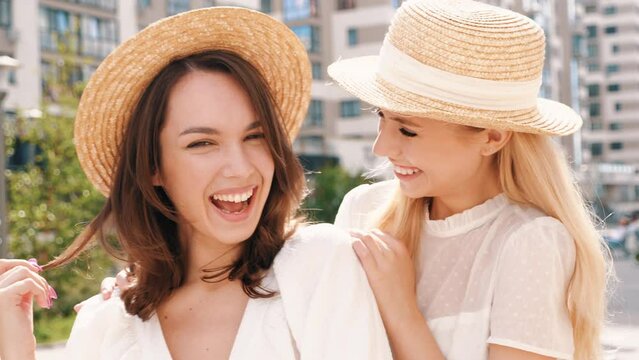 Two young beautiful smiling hipster female in trendy summer white dress clothes and straw hats. Sexy carefree women walking in street. Positive models having fun, hugging and laughing 