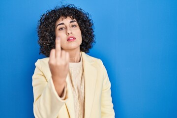Young brunette woman with curly hair standing over blue background showing middle finger, impolite...