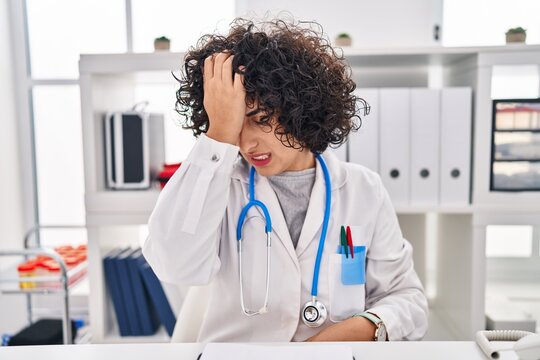 Young brunette woman with curly hair wearing doctor uniform and stethoscope surprised with hand on head for mistake, remember error. forgot, bad memory concept.