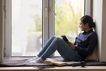 Young Indian woman holds modern smartphone staring out window resting seated on windowsill, spend...