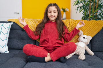 Adorable hispanic girl doing yoga exercise sitting on sofa at home
