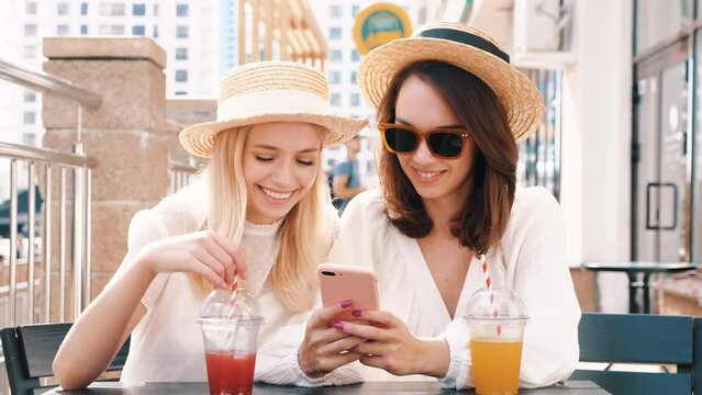Two young beautiful smiling hipster female in trendy summer white dress and hats. Sexy carefree women drinking fresh vegetable cocktail smoothie drink in plastic cup with straw. Using phone apps