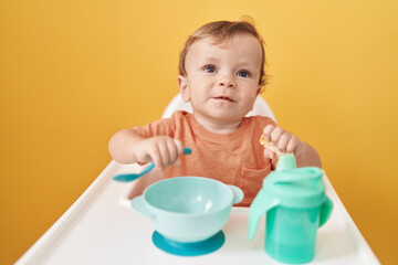 Adorable blond toddler sitting on highchair eating snack over isolated yellow background