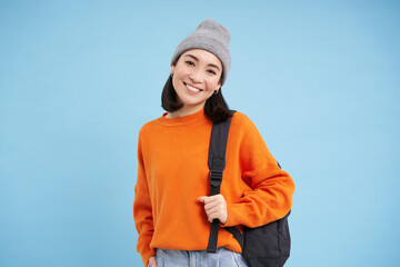 Stylish young asian woman in warm hat, walking with backpack, going somewhere with bag, standing...