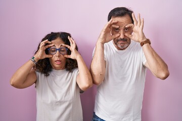 Middle age hispanic couple together over pink background trying to open eyes with fingers, sleepy and tired for morning fatigue