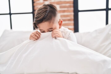 Fototapeta na wymiar Adorable hispanic boy sitting on bed covering face with bedsheet at bedroom
