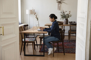 Attractive millennial Indian business woman sit at table, use laptop, create new fresh idea, type business plans text, thinks about finding solution, do remote works, learn new application or software